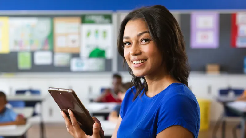 Smiling teacher holding a tablet in her classroom, ready to start an online professional development course