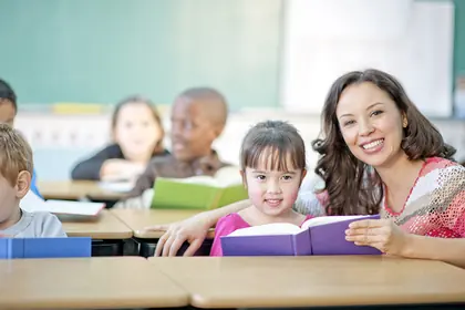 Smiling teacher sitting beside a young student holding a book in a classroom